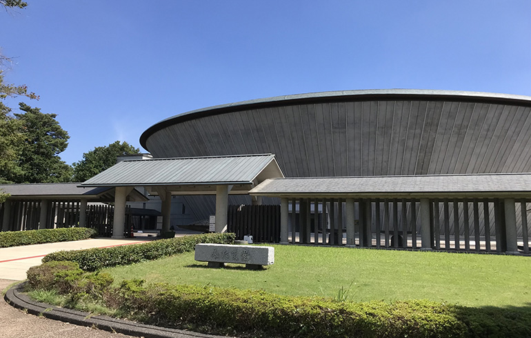 Curved, circular museum building with a dark wooden exterior and a covered entrance on a sunny day, lawn in the foreground.