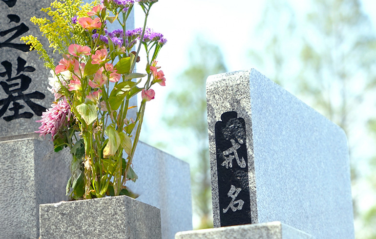 Stone grave marker with a bouquet of pink and yellow flowers laid at the base in a cemetery scene.