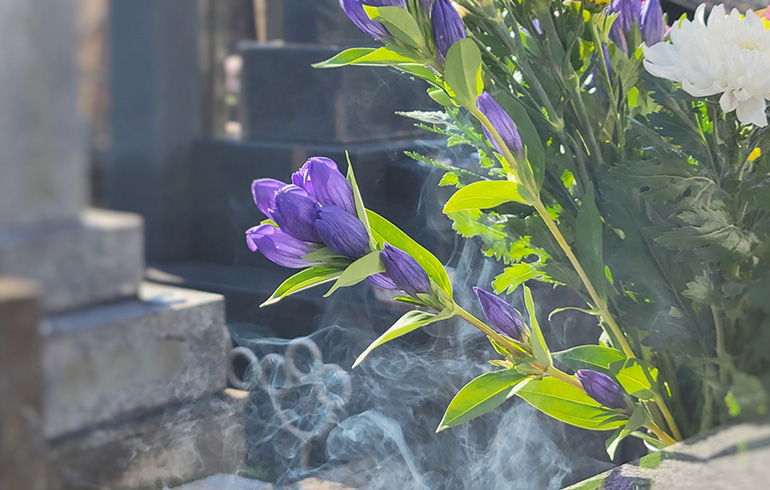 Purple flowers in a bouquet with green leaves placed at a cemetery, with wisps of incense smoke rising around a gravestone.
