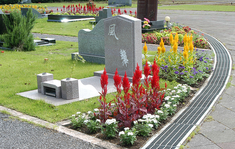 Gray tombstone with engraved characters set in a landscaped cemetery, surrounded by red, yellow, and white flowers and a curved metal grate path.