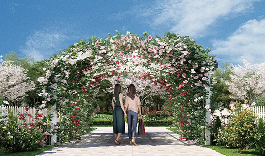 Two women walk through a white floral arch covered in pink and red roses in a sunny garden path.
