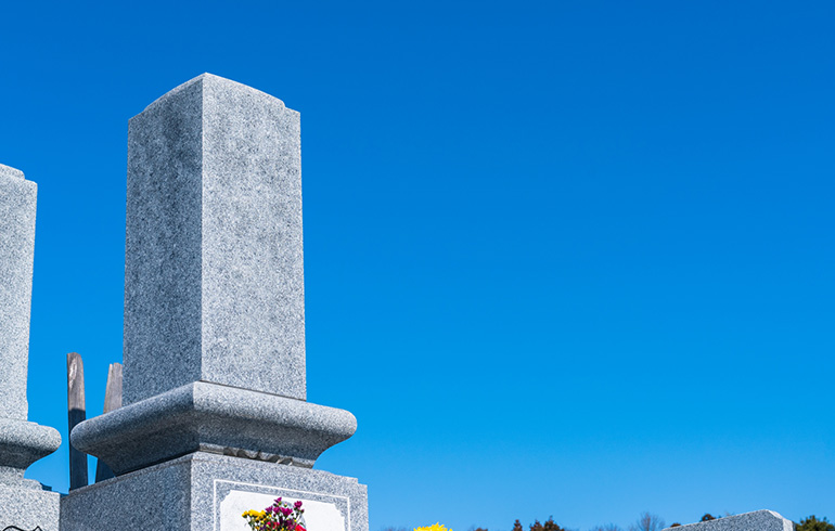 Gray granite obelisks in a cemetery with a small flower arrangement at the base against a clear blue sky.