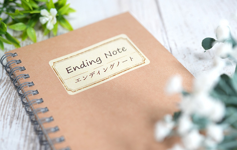 Brown spiral notebook labeled 'Ending Note' on a light wooden surface with green and white flowers nearby.